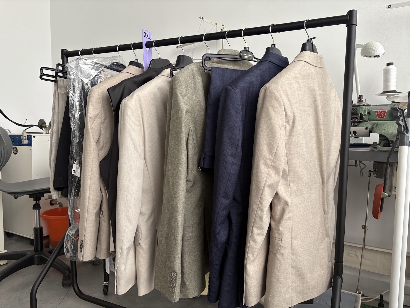 A rail of altered suit jackets in beige and navy beside an overlock machine in the Craft Tailors studio